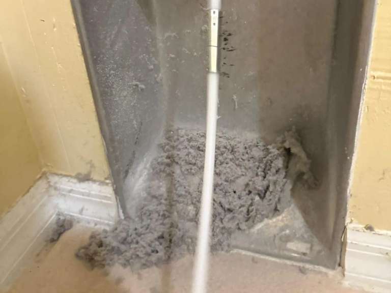 Una varilla blanca de limpieza que baja a la caja de pared del ducto de la secadora, llena de pelusa gris